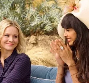 Duas mulheres sorriem ao ar livre. Uma loira em xadrez e outra de chapéu grande com franja, rindo e batendo palmas. Cena alegre e ensolarada.