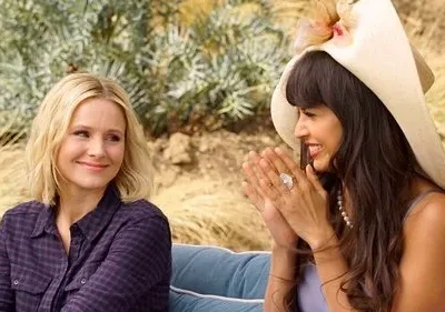 Duas mulheres sorriem ao ar livre. Uma loira em xadrez e outra de chapéu grande com franja, rindo e batendo palmas. Cena alegre e ensolarada.