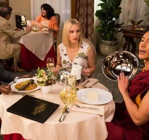 Três pessoas jantam em restaurante. Um homem de terno, uma mulher loira surpresa e um homem de robe falando, segurando tigela prateada.