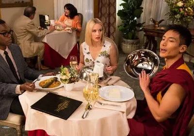 Três pessoas jantam em restaurante. Um homem de terno, uma mulher loira surpresa e um homem de robe falando, segurando tigela prateada.