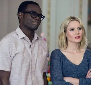 Homem negro de óculos e camisa clara sorri; mulher loira de blusa azul e braços cruzados olha pensativa. Fundo com doces.