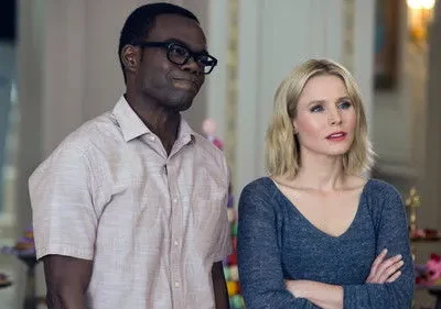 Homem negro de óculos e camisa clara sorri; mulher loira de blusa azul e braços cruzados olha pensativa. Fundo com doces.