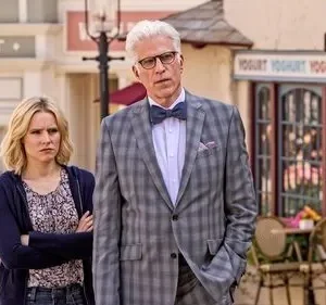 Um homem de terno xadrez e uma mulher loira com braços cruzados numa rua peculiar. Ele parece confuso, ela cética. Clima cômico.