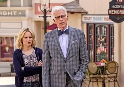 Um homem de terno xadrez e uma mulher loira com braços cruzados numa rua peculiar. Ele parece confuso, ela cética. Clima cômico.