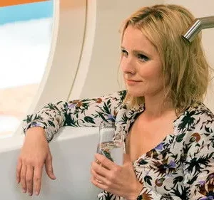 Mulher loira de vestido florido sentada em banheira, com um copo na mão, olhando pela janela. Cenário externo azul/branco.