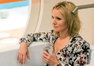 Mulher loira de vestido florido sentada em banheira, com um copo na mão, olhando pela janela. Cenário externo azul/branco.