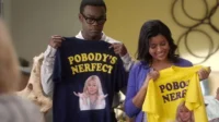 The Good Place – T01E11:Qual é a minha motivação 9 Homem e mulher seguram camisetas com POBODY'S NERFECT e loira encolhendo os ombros. Ele sério,ela sorri.