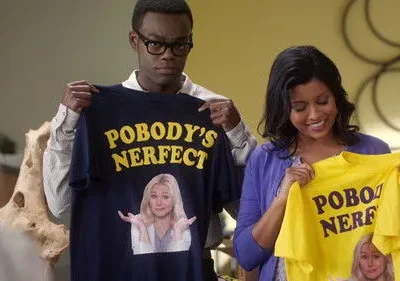 Homem e mulher seguram camisetas com POBODY'S NERFECT e loira encolhendo os ombros. Ele sério, ela sorri.