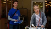 The Good Place – T01E12:Mindy St. Claire 8 Homem de camisa azul e prancheta sorri para mulher loira de capuz,com carrinho e olhar surpreso,na frente de uma loja.