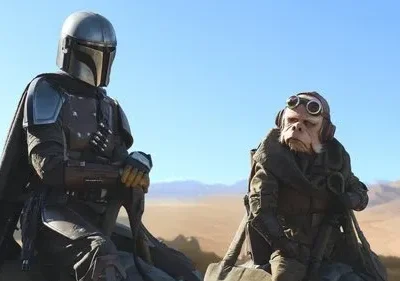 Figura blindada e alienígena com óculos em deserto árido sob céu azul. Clima de ficção científica e aventura.