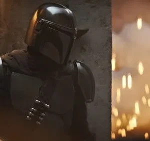 Personagem em armadura cinza-escura e capacete. Ao lado, explosão com faíscas alaranjadas e fumaça. Cena de ação intensa.