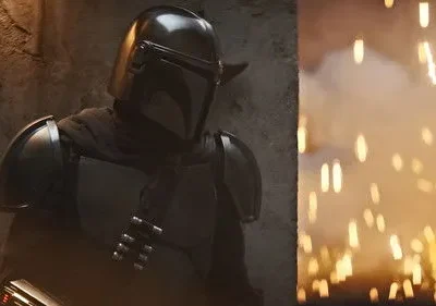 Personagem em armadura cinza-escura e capacete. Ao lado, explosão com faíscas alaranjadas e fumaça. Cena de ação intensa.