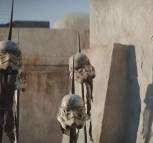 Múltiplos capacetes de ficção científica brancos e danificados espetados em postes, contra edifícios desérticos. Cena de guerra sombria.