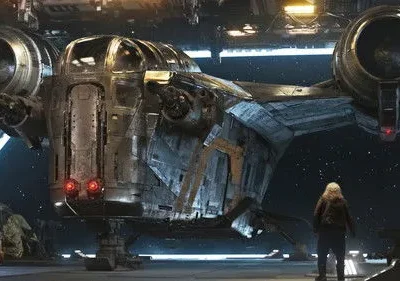 Uma figura loira de costas admira uma nave espacial robusta e desgastada em um hangar. O céu estrelado aparece ao fundo.