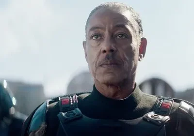 Homem de meia-idade, com bigode e olhar sério, veste uniforme escuro com armadura. Fundo de céu claro e edificações futuristas.