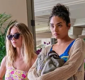 Duas mulheres em um resort. Uma loira de óculos e top floral; a outra com coque e expressão tensa, segurando uma bolsa.