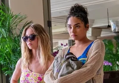 Duas mulheres em um resort. Uma loira de óculos e top floral; a outra com coque e expressão tensa, segurando uma bolsa.