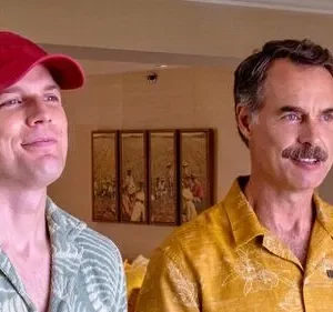 Dois homens sorrindo, em camisas havaianas. O da esquerda usa boné vermelho, o da direita tem bigode e camisa amarela. Clima descontraído.