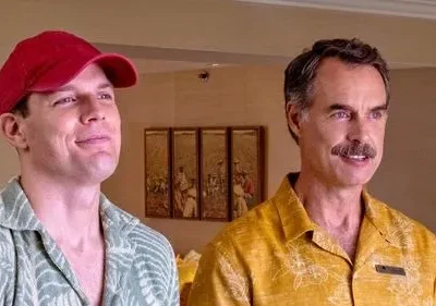 Dois homens sorrindo, em camisas havaianas. O da esquerda usa boné vermelho, o da direita tem bigode e camisa amarela. Clima descontraído.