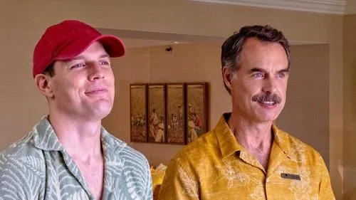 Dois homens sorrindo,em camisas havaianas. O da esquerda usa boné vermelho,o da direita tem bigode e camisa amarela. Clima descontraído.