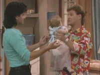 Homem de camisa estampada segura bebê, interagindo com mulher de blusa azul clara. Cenário doméstico familiar e descontraído.