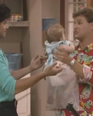 Homem de camisa estampada segura bebê, interagindo com mulher de blusa azul clara. Cenário doméstico familiar e descontraído.