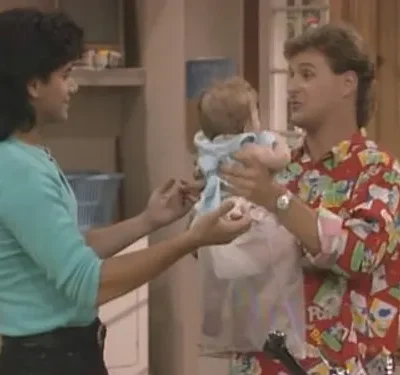 Homem de camisa estampada segura bebê, interagindo com mulher de blusa azul clara. Cenário doméstico familiar e descontraído.