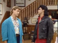 Dois homens com visuais vibrantes dos anos 90, um de blazer azul, outro de jaqueta de couro preta, em uma casa com escada.