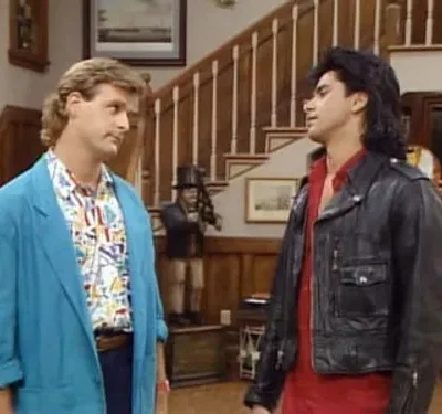 Dois homens com visuais vibrantes dos anos 90, um de blazer azul, outro de jaqueta de couro preta, em uma casa com escada.