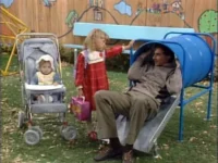 Um homem sorridente sai de um túnel azul no playground. Uma menina de vestido vermelho e um bebê no carrinho estão ao lado.