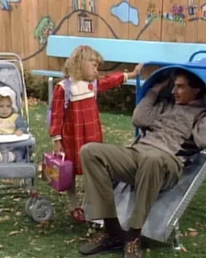 Um homem sorridente sai de um túnel azul no playground. Uma menina de vestido vermelho e um bebê no carrinho estão ao lado.