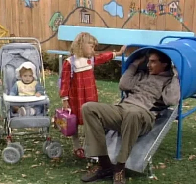 Um homem sorridente sai de um túnel azul no playground. Uma menina de vestido vermelho e um bebê no carrinho estão ao lado.