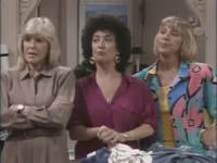 Três mulheres em ambiente interno. Loira de bege com braços cruzados, morena de roxo e loira com camisa colorida abstrata. Expressões variadas.