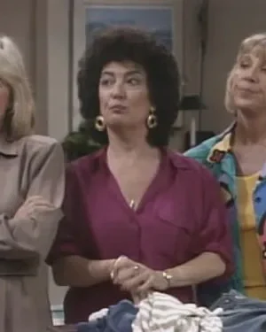 Três mulheres em ambiente interno. Loira de bege com braços cruzados, morena de roxo e loira com camisa colorida abstrata. Expressões variadas.