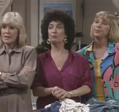 Três mulheres em ambiente interno. Loira de bege com braços cruzados, morena de roxo e loira com camisa colorida abstrata. Expressões variadas.