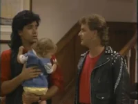 Homem de jaqueta rocker com tachas olha para homem de mullet surpreso segurando um bebê. Cena de comédia familiar em casa.