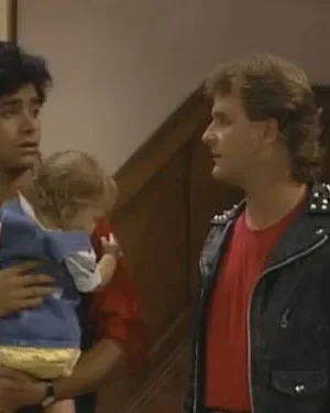 Homem de jaqueta rocker com tachas olha para homem de mullet surpreso segurando um bebê. Cena de comédia familiar em casa.