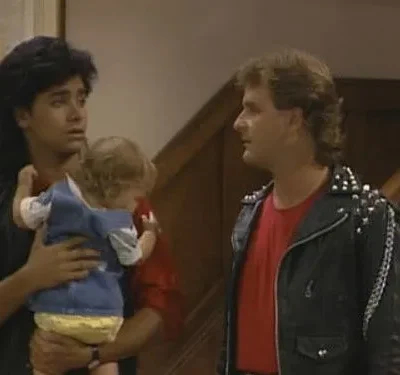 Homem de jaqueta rocker com tachas olha para homem de mullet surpreso segurando um bebê. Cena de comédia familiar em casa.