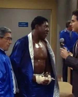 Um repórter de terno entrevista um boxeador em robe azul e ataduras, com um treinador ao lado, em um vestiário.