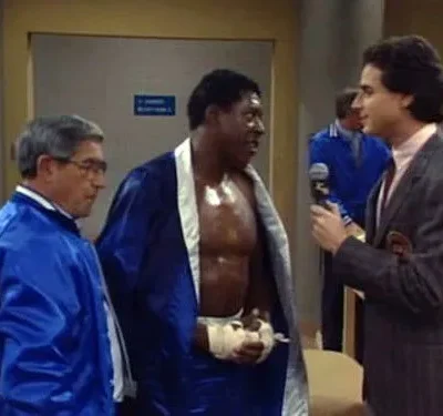 Um repórter de terno entrevista um boxeador em robe azul e ataduras, com um treinador ao lado, em um vestiário.