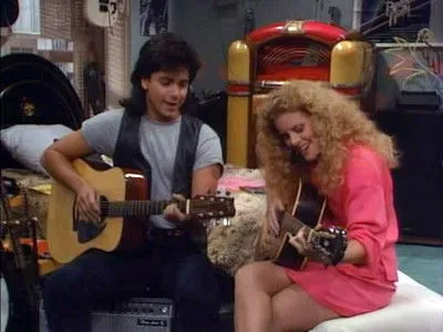 Homem de mullet e mulher de cachos loiros sorriem tocando violão em quarto musical com jukebox. Ambiente dos anos 80.