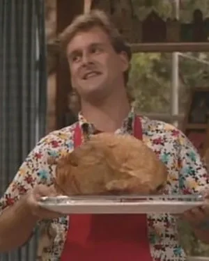 Homem sorridente com mullet, camisa florida e avental vermelho, segurando um peru assado em uma bandeja, em ambiente doméstico.
