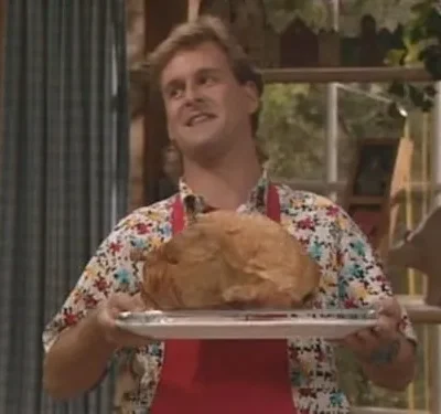 Homem sorridente com mullet, camisa florida e avental vermelho, segurando um peru assado em uma bandeja, em ambiente doméstico.