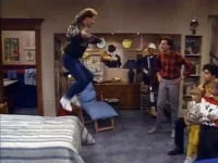 Um homem salta sobre a cama em uma sala de estar retrô. Várias pessoas, adultos e crianças, observam a cena animada.