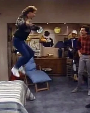 Um homem salta sobre a cama em uma sala de estar retrô. Várias pessoas, adultos e crianças, observam a cena animada.