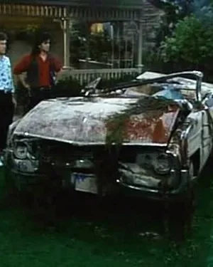 Família olha um carro conversível totalmente destruído e coberto de detritos em um gramado. Atmosfera de choque e surpresa.