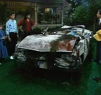 Família olha um carro conversível totalmente destruído e coberto de detritos em um gramado. Atmosfera de choque e surpresa.