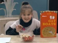 Garota sorridente de suéter come tigela de cereal com frutas. Uma caixa de OAT BOATS está na mesa de um ambiente caseiro.
