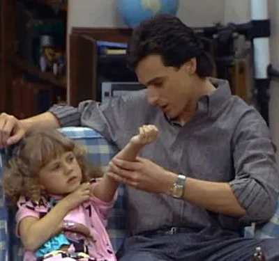 Homem e menina em sofá xadrez. Ele examina o braço dela com carinho. Clima acolhedor e de cuidado.