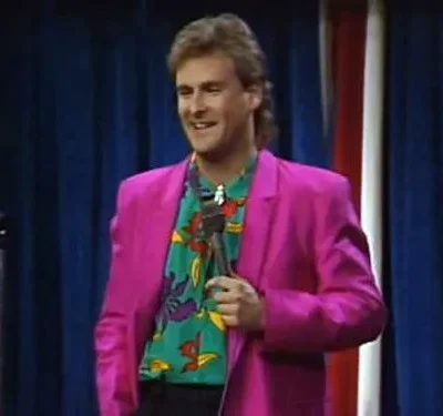 Homem sorridente em blazer pink e camisa colorida segura microfone em palco com cortinas azuis e listras. Estilo anos 80/90.